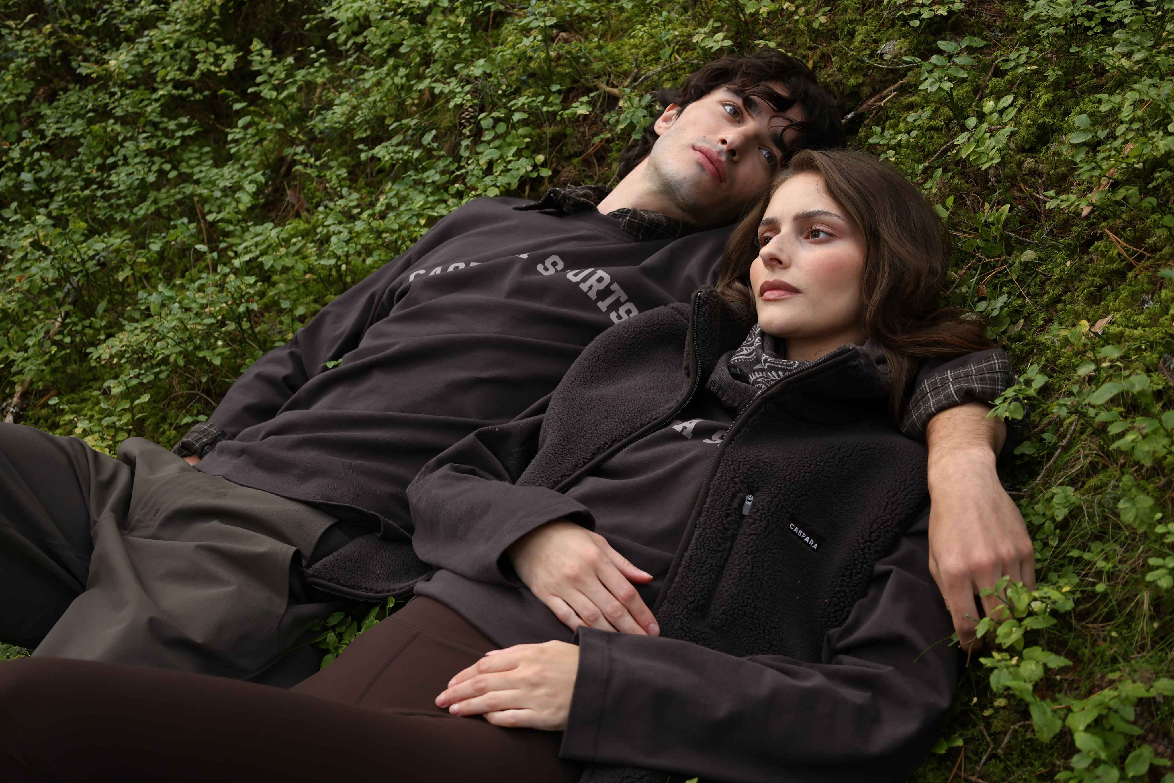 Man and woman lying on lush forest moss wearing espresso CASPARA fleece vest and top.